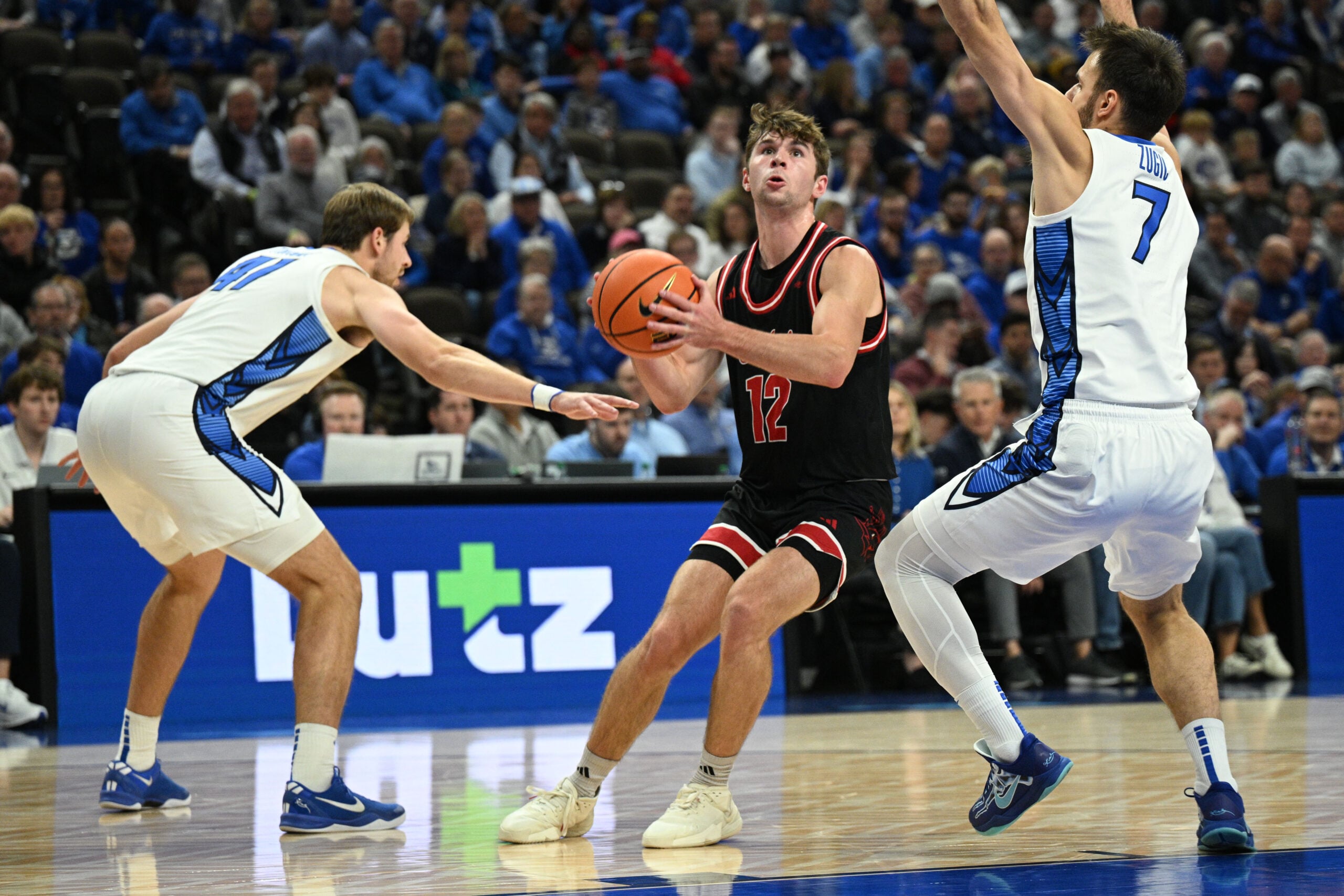 Nov 5, 2025; Omaha, Nebraska, USA; South Dakota Coyotes guard Isaac Bruns (12) drives against Creighton Bluejays forward Isaac Traudt (41) and Fedor Zugic (7) during the first half at CHI Health Center Omaha. Mandatory Credit: Steven Branscombe-Imagn Images
